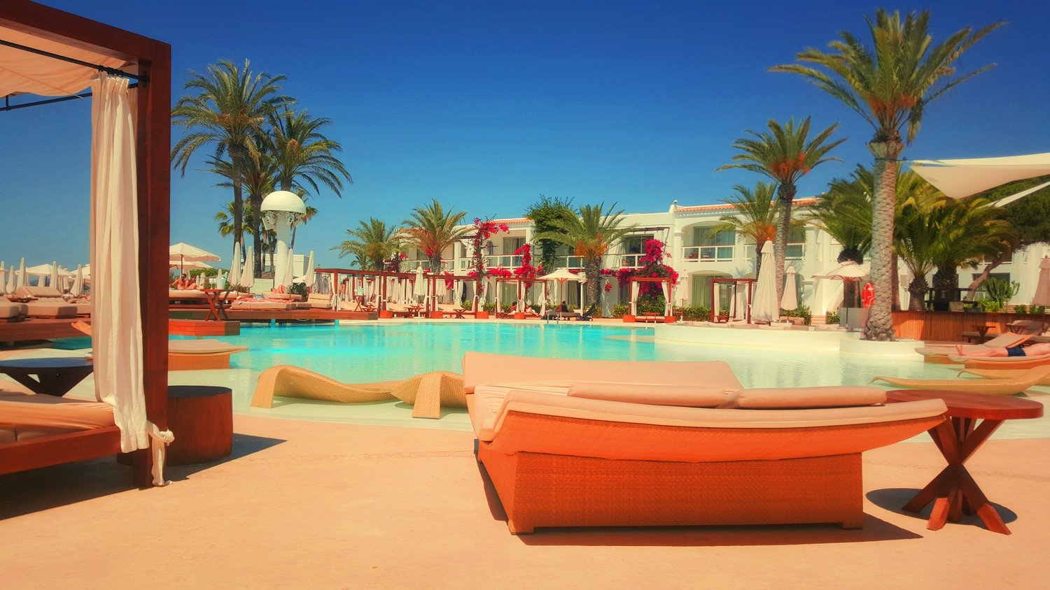 Sun loungers beside a turquoise resort pool lined with palm trees and white villas